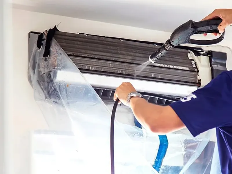 Technicien qui nettoie au jet d'eau le serpentin d'une thermopompe murale, protégée par une housse de plastique